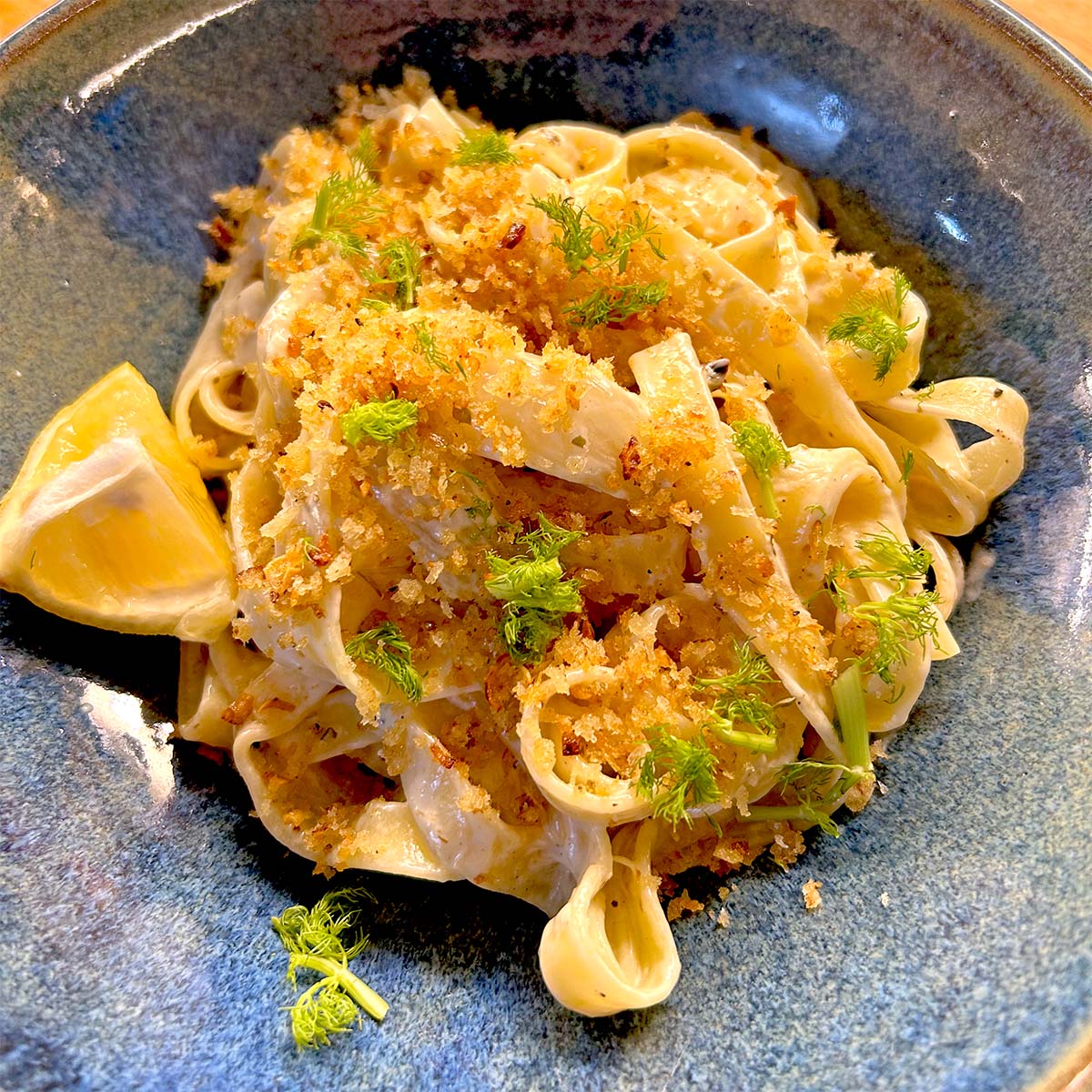 A bowl of fettuccine pasta topped with golden breadcrumbs and fresh herbs, garnished with fennel fronds and served with a lemon wedge on the side.