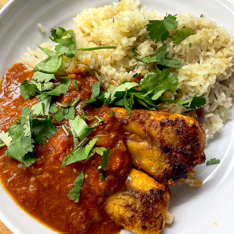 A plate of chicken curry with rich red sauce, served alongside seasoned rice and garnished with fresh coriander leaves.