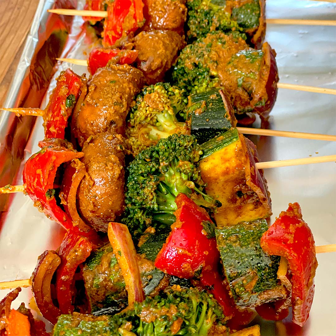 Vegetable skewers with mushrooms, broccoli, courgette, red peppers, and onions covered in seasoning, arranged on a foil-lined tray.