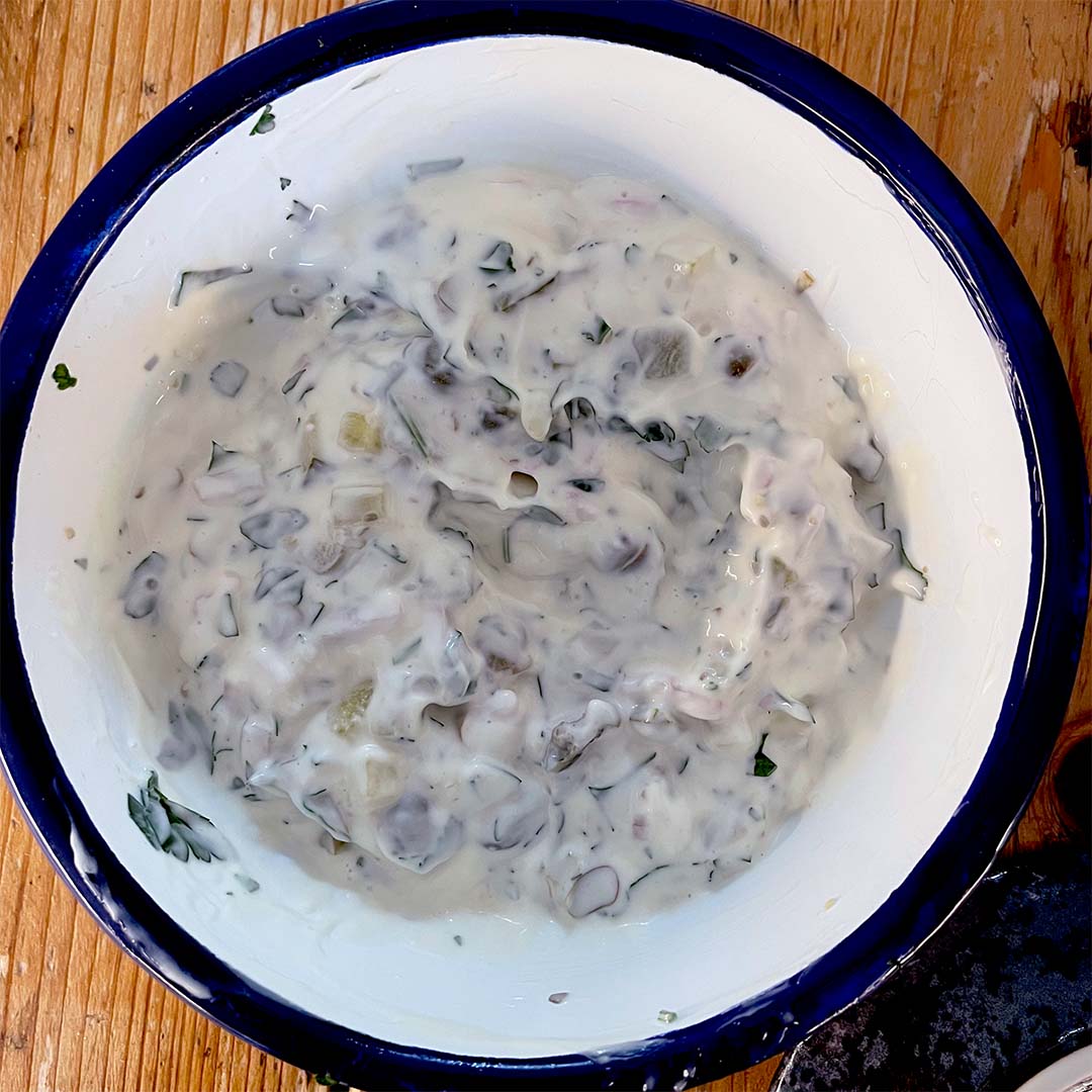 A white bowl with a blue rim filled with a creamy mixture containing chopped herbs and vegetables, sitting on a wooden surface.