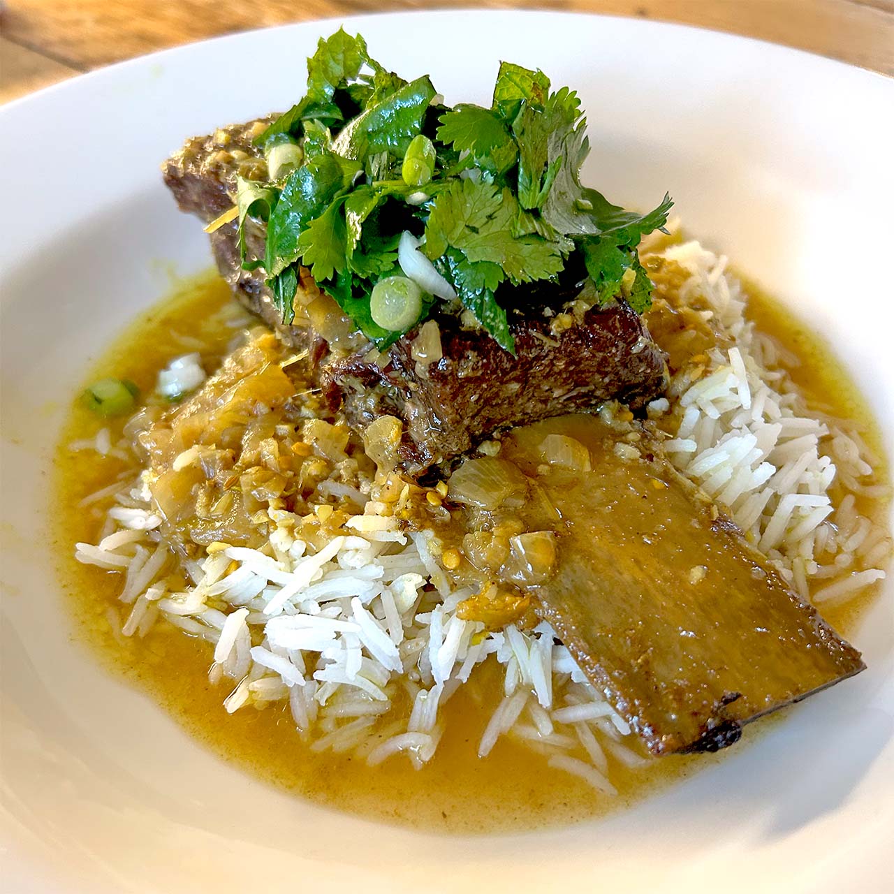 A braised beef short rib served over white rice, topped with fresh coriander and chopped spring onions, with a golden sauce poured around the dish on a white plate.