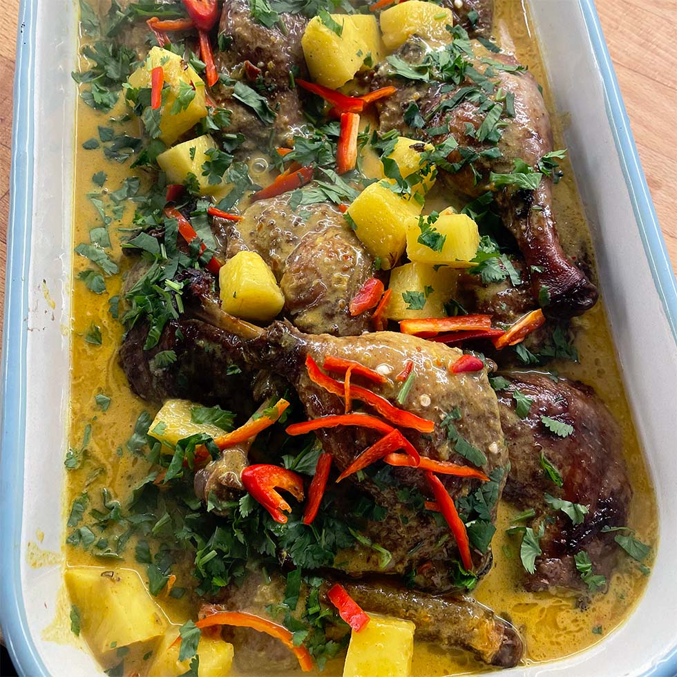 A roasting dish filled with roasted chicken pieces in a yellow curry sauce, garnished with pineapple chunks, chopped coriander, and sliced red chilli peppers.