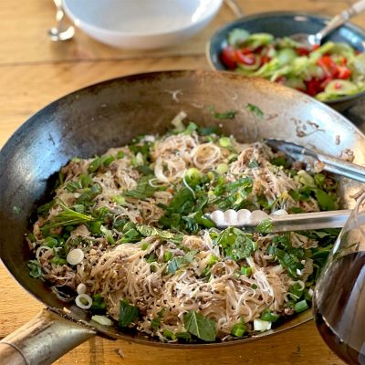 A large pan filled with cooked rice noodles, chopped spring onions, herbs, and sesame seeds, with metal tongs resting on the side. In the background, there is a bowl of mixed salad with tomatoes and cucumbers.