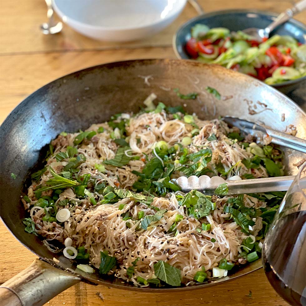 A large pan filled with cooked rice noodles, chopped spring onions, herbs, and sesame seeds, with metal tongs resting on the side. In the background, there is a bowl of mixed salad with tomatoes and cucumbers.