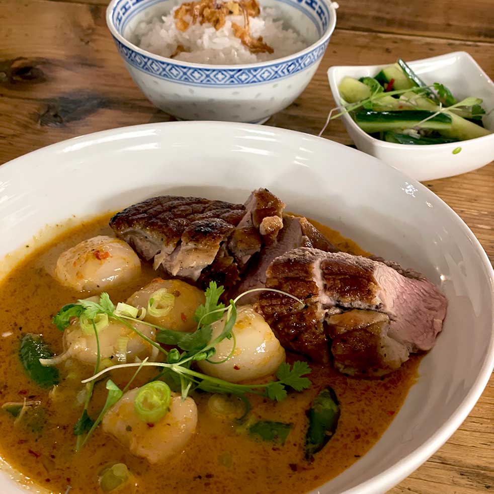 A bowl of red curry with sliced duck, round vegetables, and herbs, served with a side of white rice in a patterned bowl and a small dish of cucumber salad on a wooden table.