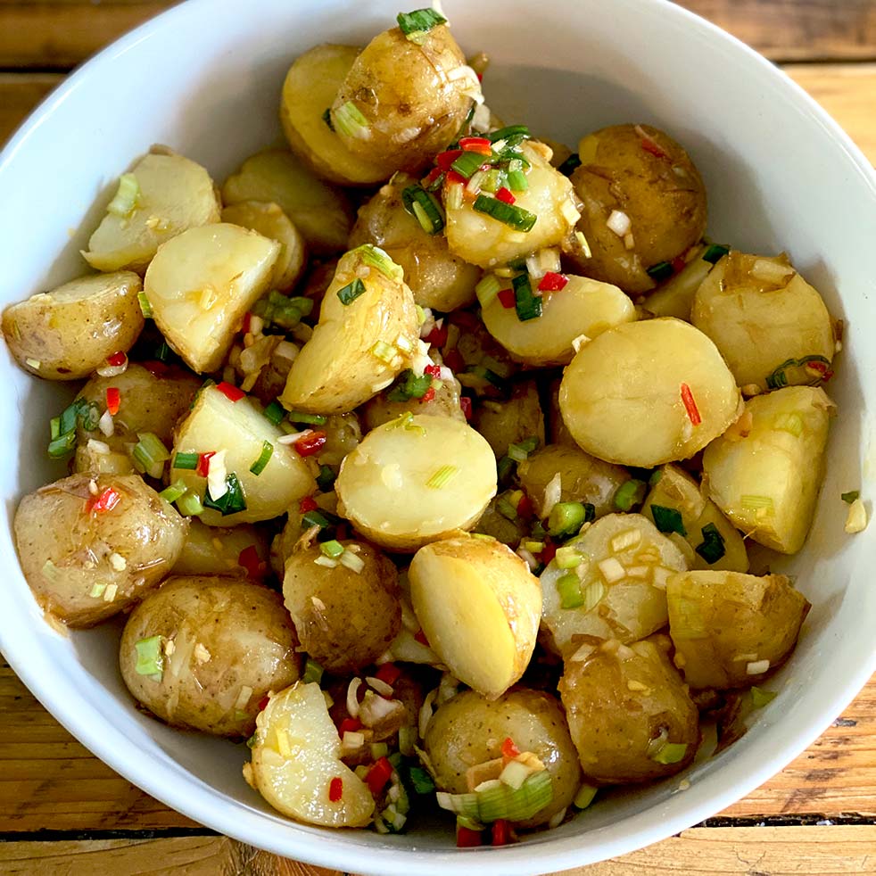 A white bowl filled with halved baby potatoes mixed with chopped spring onions, red chilli, and herbs, placed on a wooden surface.