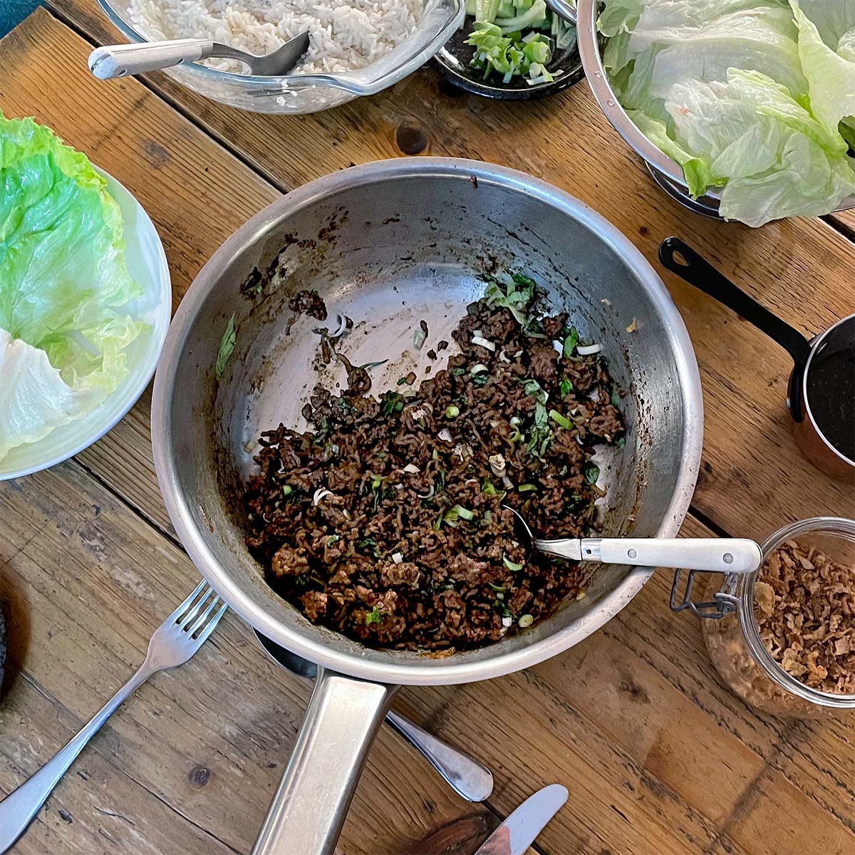 A metal pan with cooked minced meat and chopped spring onions sits on a wooden table, surrounded by lettuce leaves, a bowl of rice, cutlery, and small dishes with various toppings.
