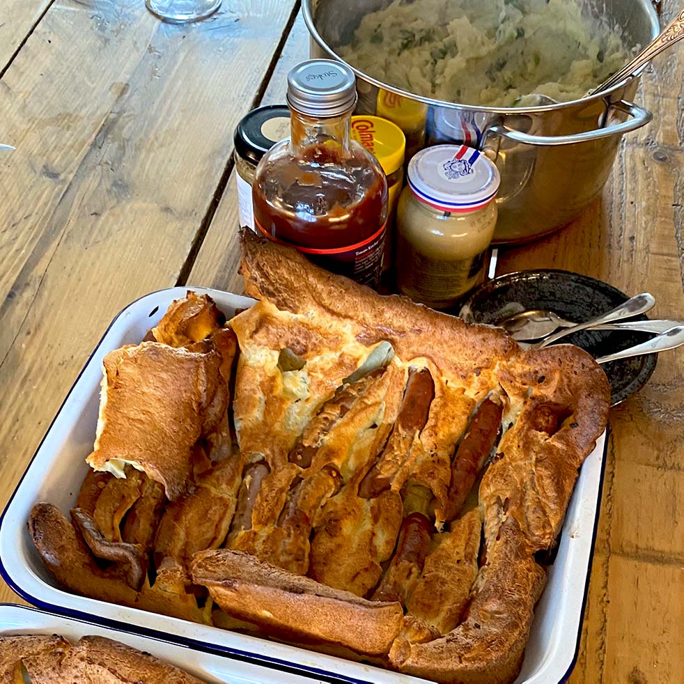 A tray of toad in the hole (sausages baked in Yorkshire pudding batter) sits on a wooden table next to a pot of mashed potato, jars of condiments, and some serving utensils.