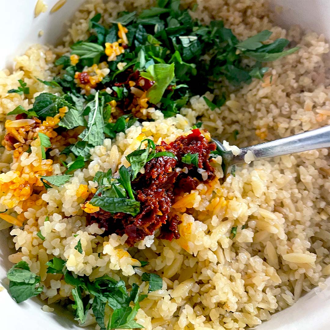 A close-up of cooked rice mixed with fresh chopped herbs and a spoonful of red sauce, stirred together in a white bowl with a metal spoon.