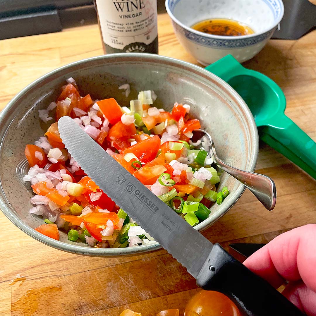 A hand holds a knife above a bowl of chopped tomatoes, onions, and green peppers. Nearby are a spoon, a green citrus squeezer, a bottle of white wine vinegar, and a bowl with a brown liquid.