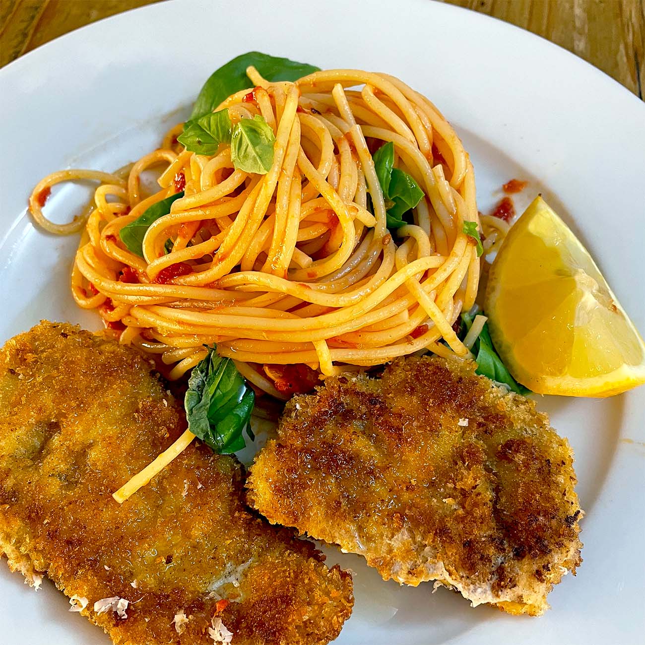 A plate with a serving of spaghetti in tomato sauce, garnished with basil leaves, two pieces of breaded fried meat, and a lemon wedge on the side.