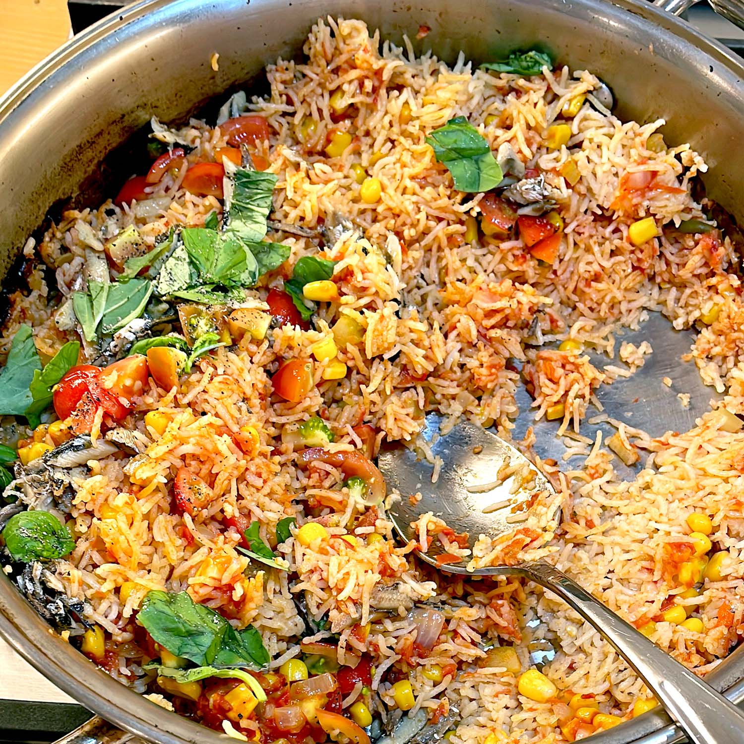 A large metal pan filled with mixed rice, sweetcorn, tomatoes, leafy herbs, and small pieces of fish. A serving spoon rests in the rice, which appears freshly cooked and partially served.
