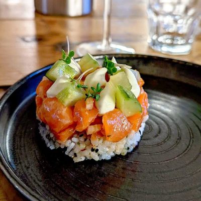 A round serving of sushi rice topped with diced salmon, avocado slices, diced cucumber, and microgreens, presented on a black plate with a glass of water in the background.