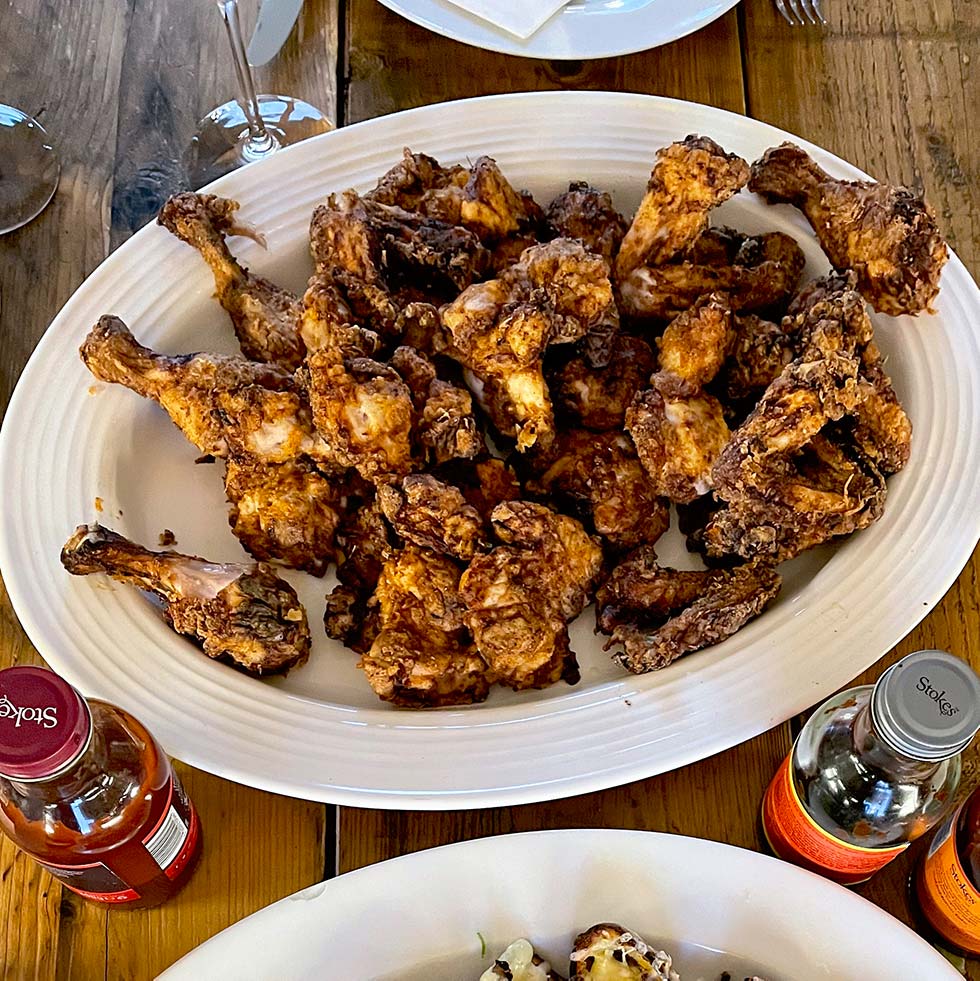 A large oval platter filled with crispy fried chicken drumsticks sits on a wooden table, surrounded by bottles of sauce and part of another dish visible at the bottom edge.