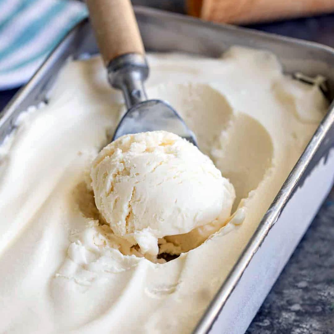 A metal ice cream scoop with a wooden handle is scooping vanilla ice cream from a rectangular metal container filled with smooth, creamy ice cream.