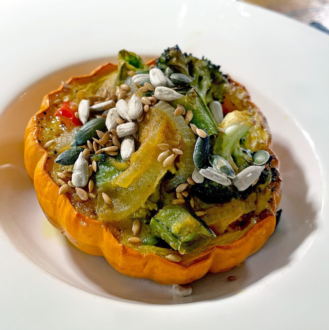 A baked orange squash half filled with roasted vegetables, topped with sunflower, pumpkin, and sesame seeds, served on a white plate.