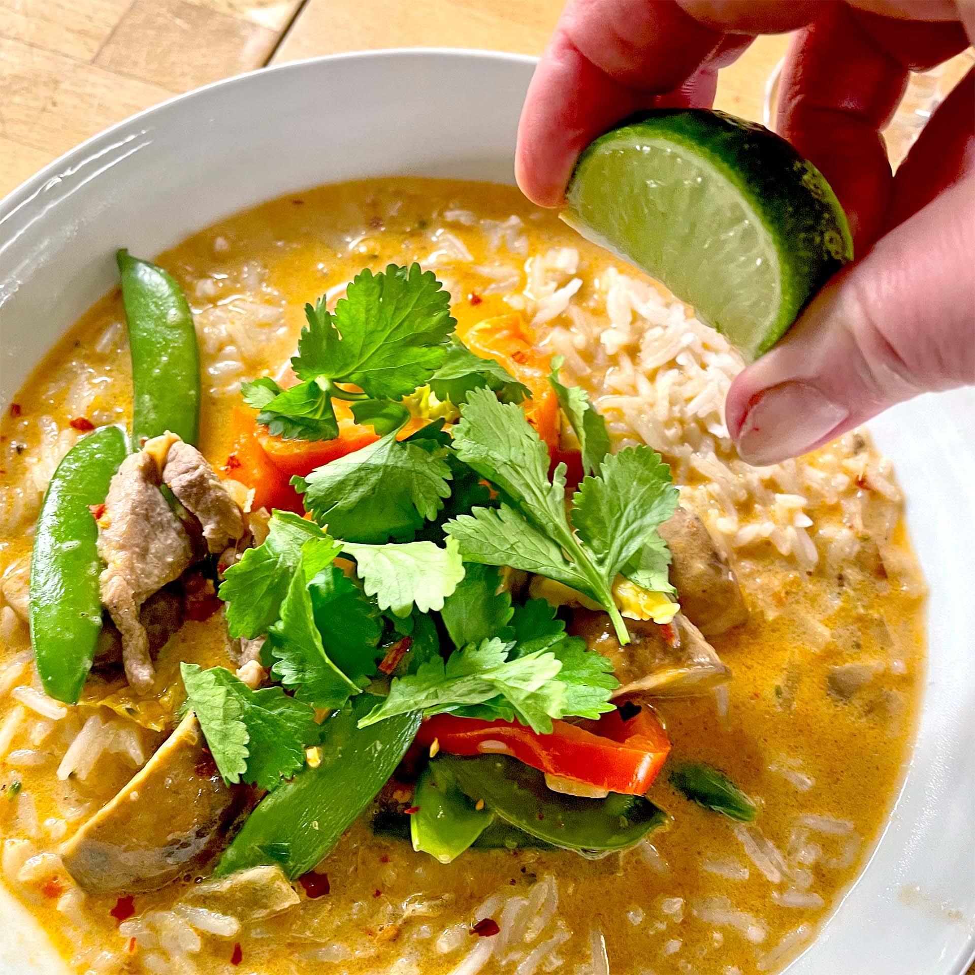 A hand squeezes a lime wedge over a bowl of rice and curry with vegetables, garnished with fresh coriander.