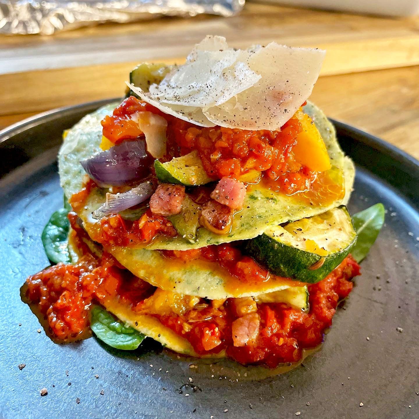 A stack of courgette slices layered with tomato sauce, diced vegetables, and pancetta, topped with grated cheese and black pepper, served on a dark plate with spinach leaves.