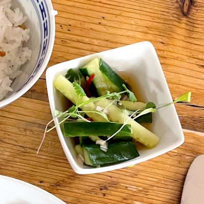 A small white bowl with cucumber slices, green herbs, and microgreens sits on a wooden table next to a bowl of white rice.