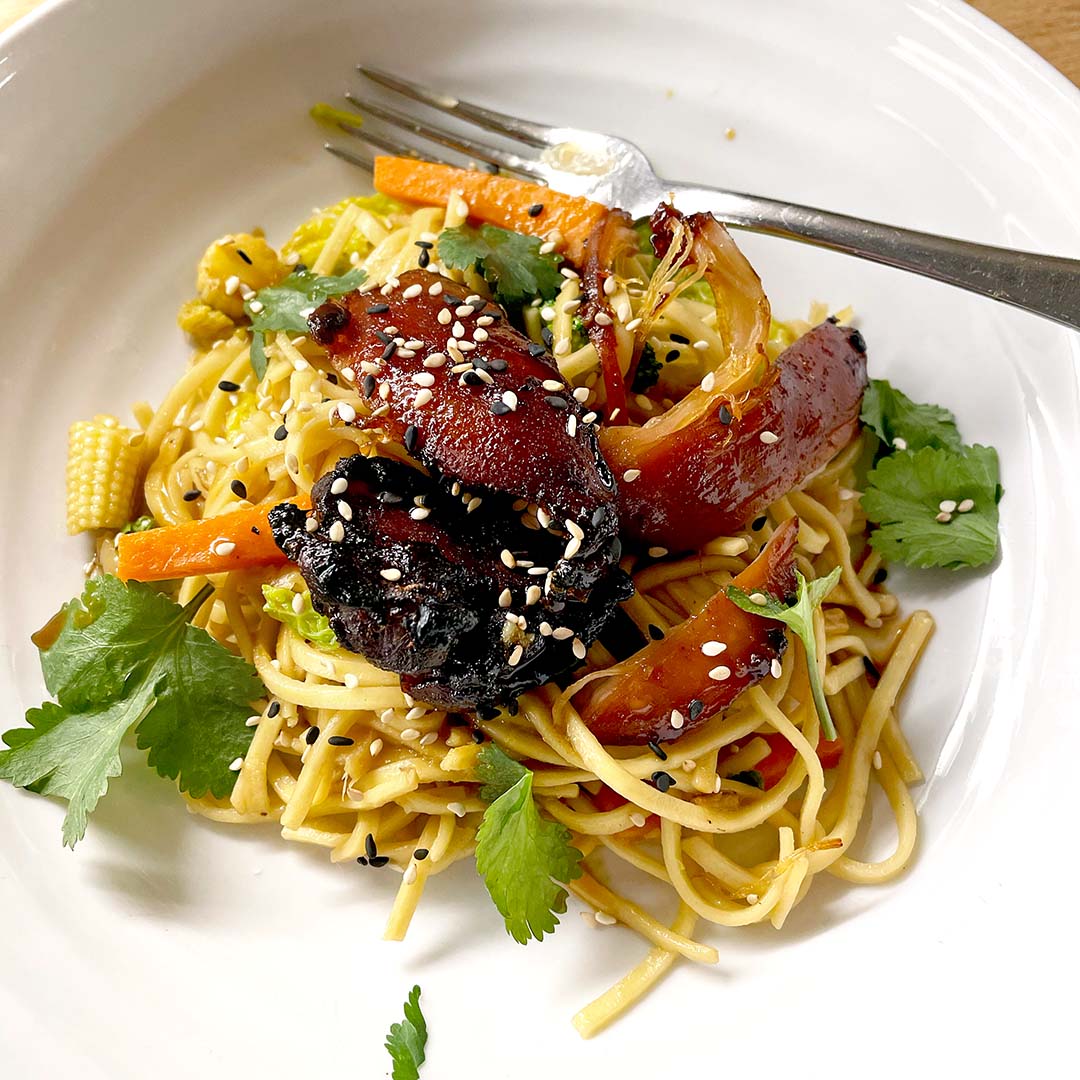 A white plate with stir-fried noodles, glazed vegetables, charred pieces of vegan mock duck, baby corn, carrots, fresh coriander leaves, and sesame seeds, with a fork resting on the edge.