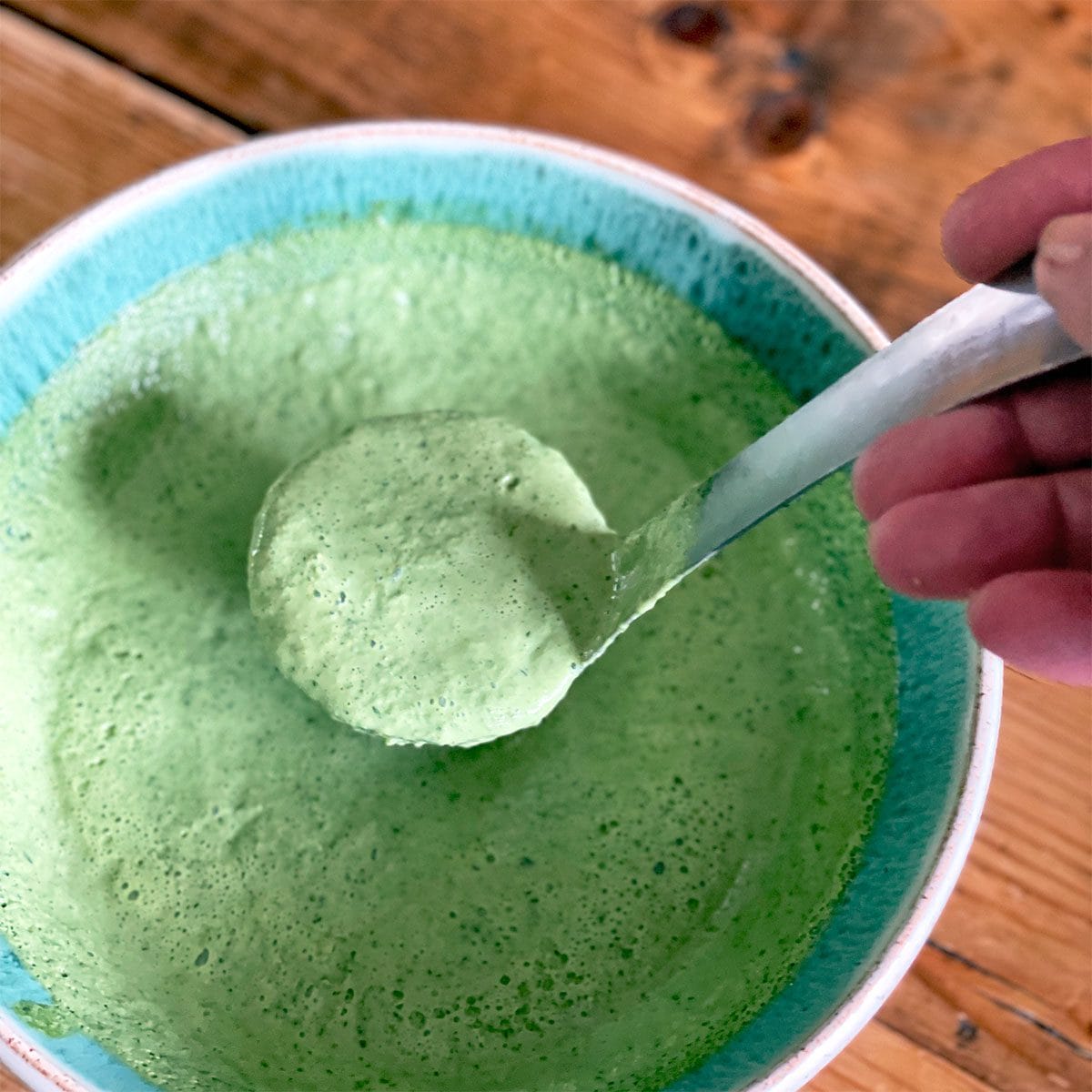 A hand holds a spoon scooping thick, green sauce or dressing from a turquoise-rimmed bowl on a wooden table. The sauce appears creamy and well-blended.