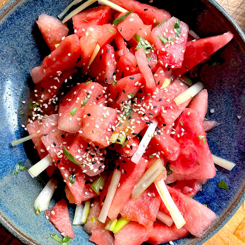 A bowl of watermelon pieces mixed with thinly sliced vegetables, topped with sesame seeds and chopped herbs, served in a blue ceramic dish.