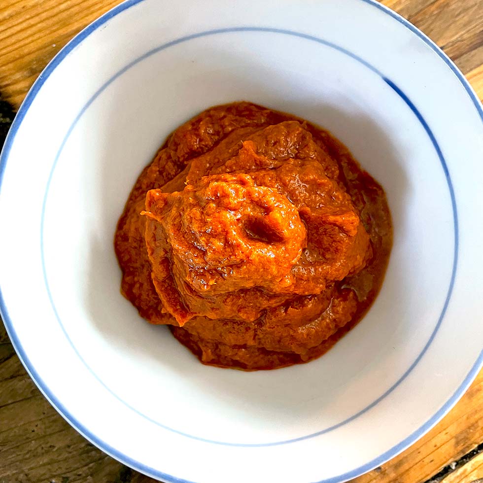 A white bowl with a blue rim filled with a thick, reddish-orange paste or sauce, placed on a wooden surface.