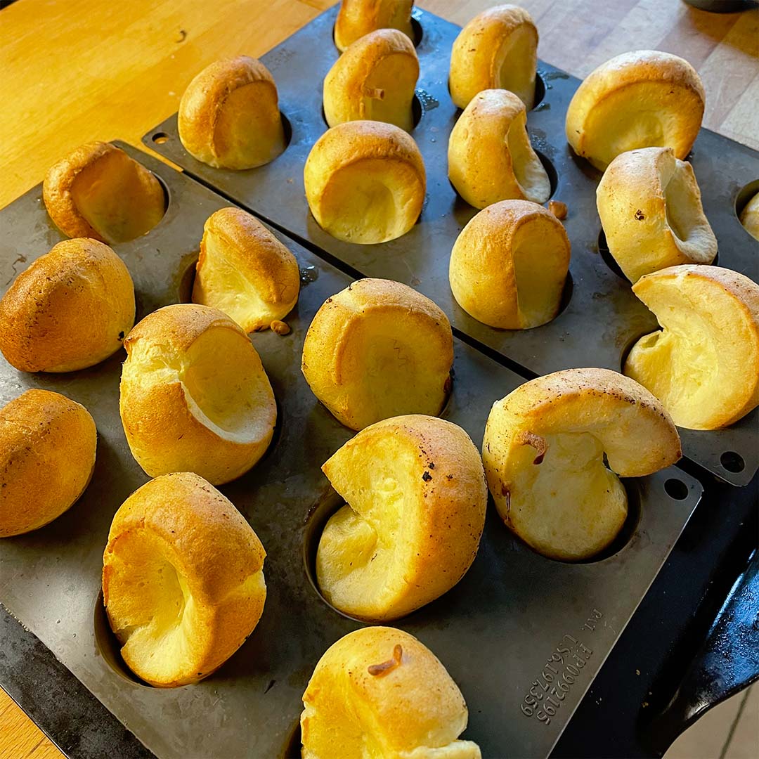Golden-brown Yorkshire puddings rise and puff out of a black muffin tray, fresh from the oven, sitting on a wooden worktop.
