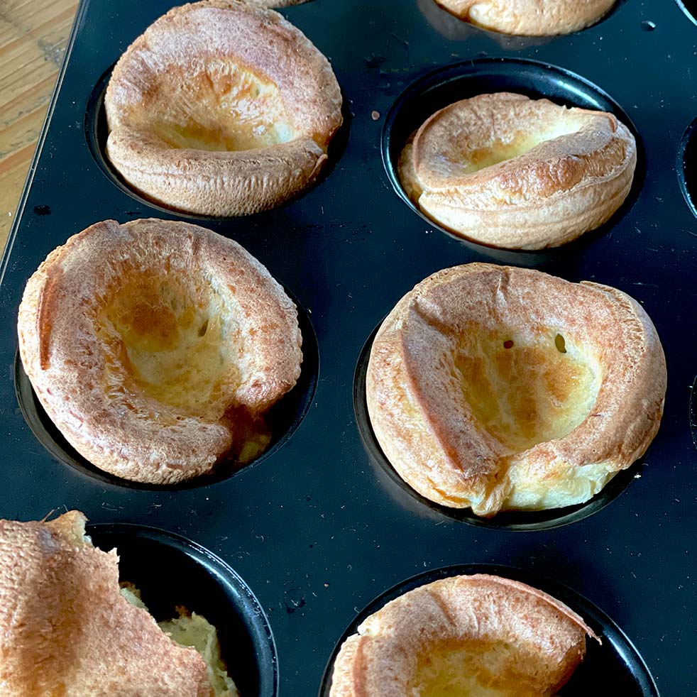 A close-up of freshly baked Yorkshire puddings in a muffin tray, showing their golden-brown, puffy edges and hollow centres.
