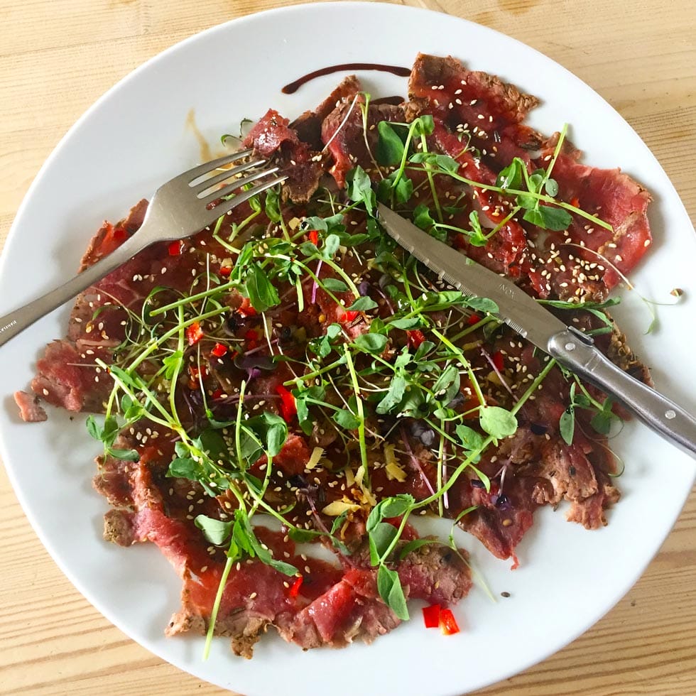A plate of thinly sliced beef carpaccio topped with microgreens, sesame seeds, and chopped red peppers, with a fork and knife resting on the plate.