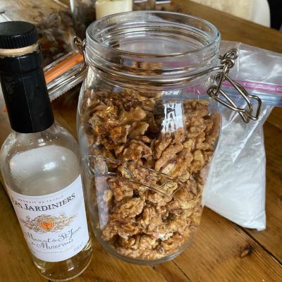 A large glass jar filled with candied walnuts sits on a wooden table next to a partially empty bottle of white wine. A plastic bag is in the background.