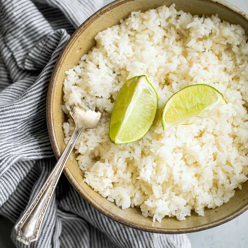A bowl of white rice topped with two lime wedges, with a silver fork resting at the side. The bowl is placed on a grey and white striped cloth.