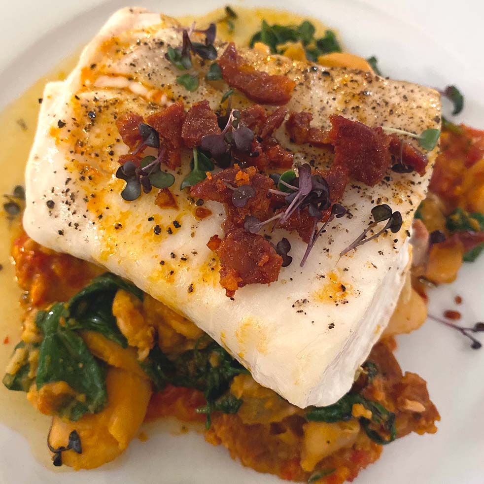 A fillet of white fish topped with microgreens and crispy bacon bits, served on a bed of wilted spinach, beans, and a tomato-based sauce on a white plate.