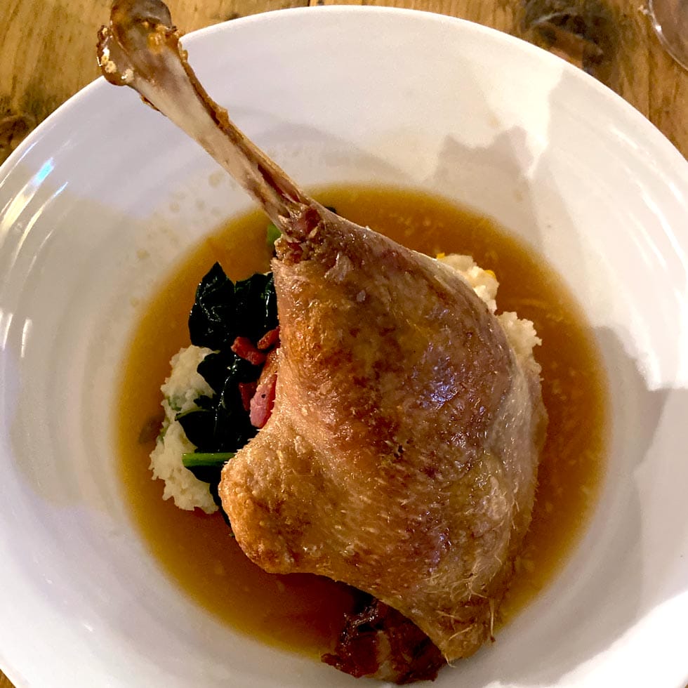 A cooked duck leg served on a bed of mashed potato and sautéed greens, surrounded by a pool of brown sauce on a white plate.