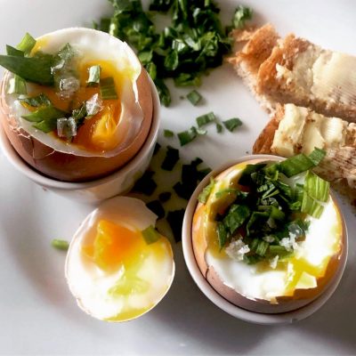 Two soft-boiled eggs in egg cups, topped with chopped spring onions and salt, with egg soldiers (buttered toast strips) and extra spring onions on a white plate. The yolks are runny and vibrant yellow.