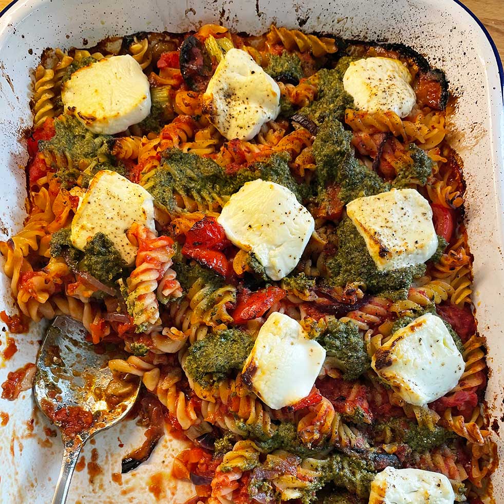 A baked pasta dish with spiral pasta, tomato sauce, dollops of green pesto, and several slices of melted white cheese on top, served in a white rectangular baking dish with a large serving spoon.