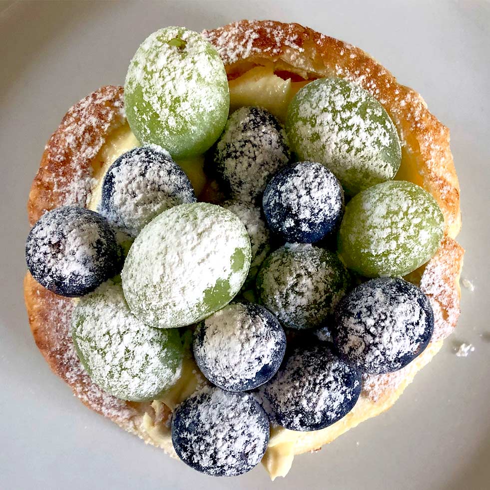 A pastry topped with green and purple grapes and dusted with icing sugar, placed on a white plate.