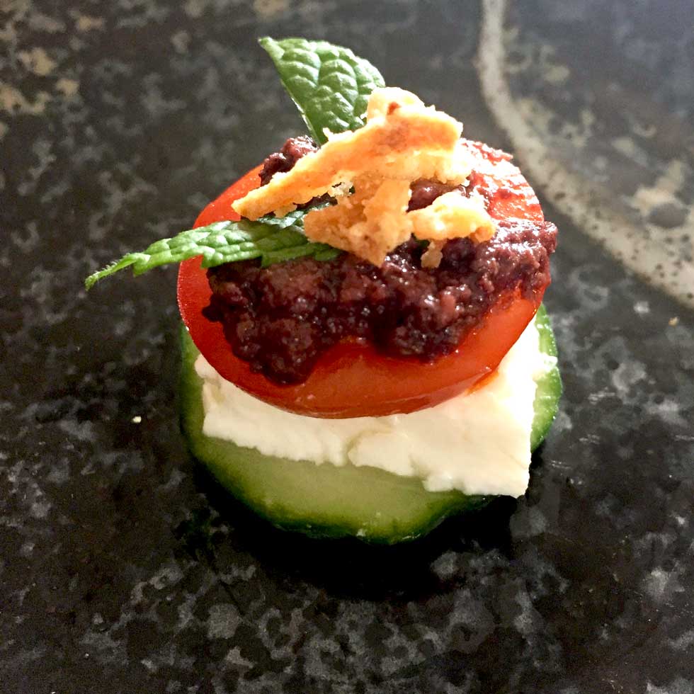 A cucumber slice topped with a layer of white cheese, a cherry tomato slice, olive tapenade, two mint leaves, and crispy fried onions, arranged on a dark, textured surface.