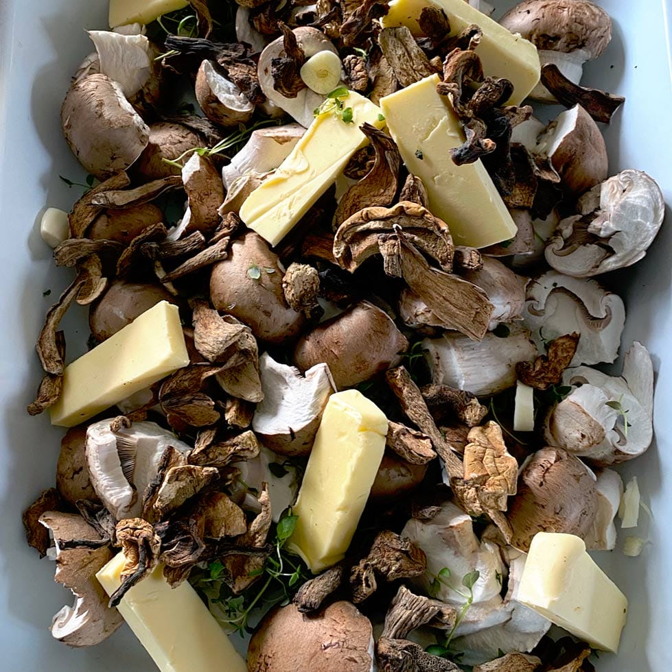 A close-up of raw mushrooms, both whole and sliced, mixed with dried mushrooms, sprigs of fresh herbs, and rectangular pieces of butter in a white dish.