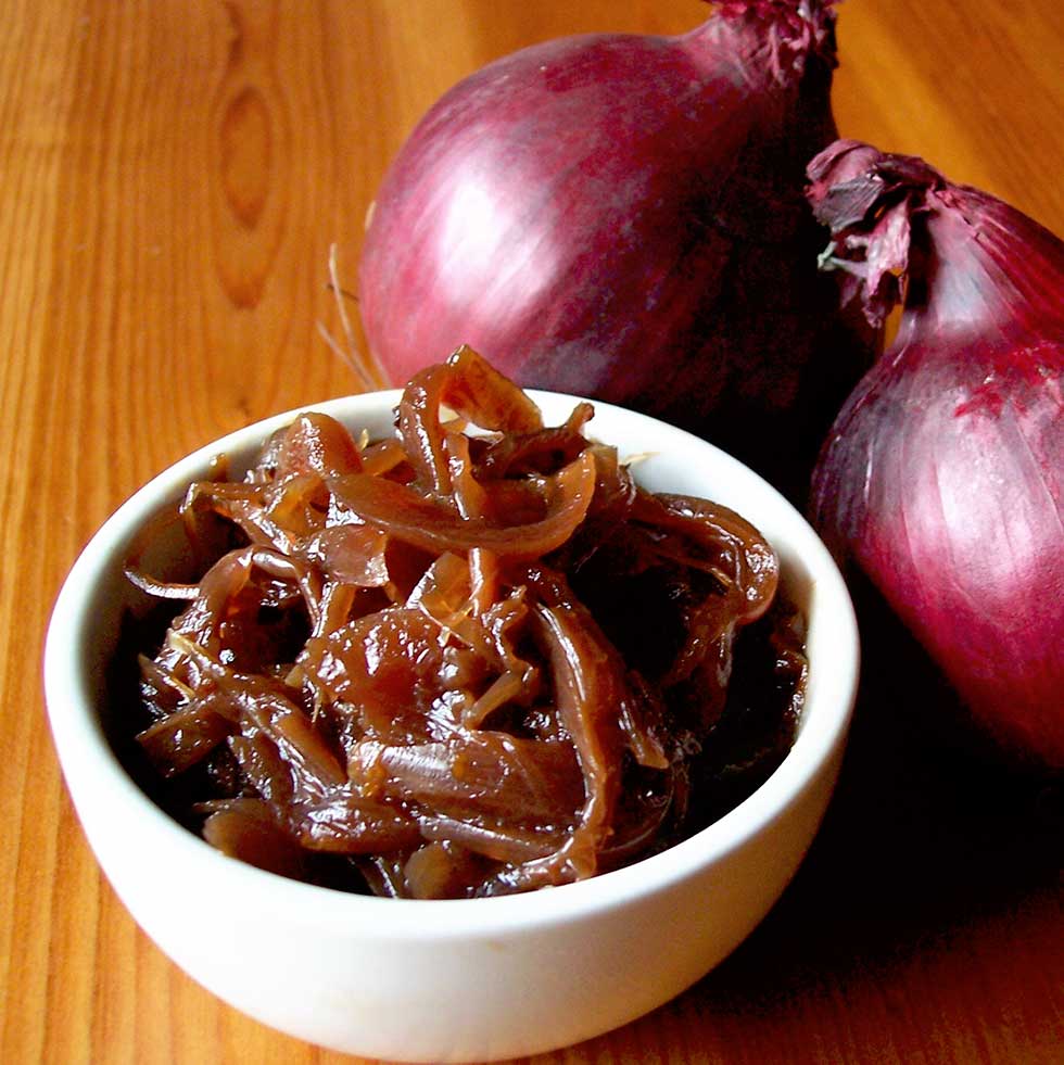 A small white bowl filled with caramelised onions sits on a wooden surface next to two whole red onions.