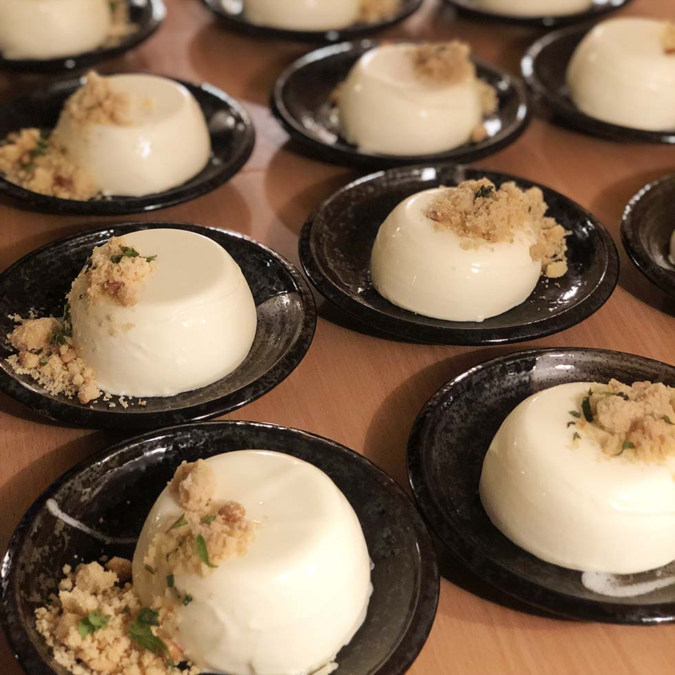 Several black plates, each with a serving of creamy white panna cotta topped with a small crumble and herbs, are arranged on a wooden surface, ready to be served.