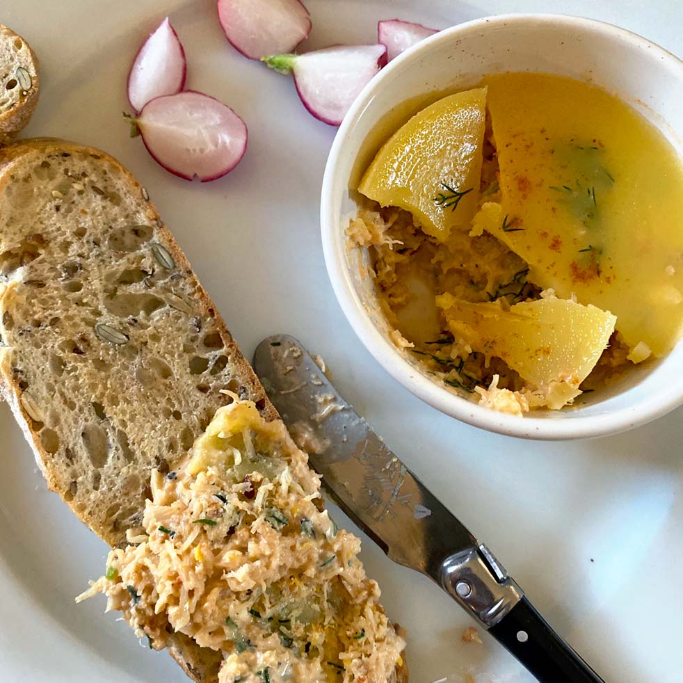A slice of multigrain bread spread with a salmon pâté sits on a plate next to a bowl of pâté topped with jelly, a butter knife, and slices of fresh radish.
