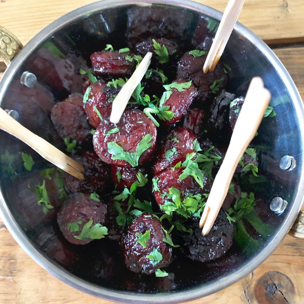 A metal bowl filled with glazed sausage slices garnished with chopped parsley, with several wooden sticks inserted for serving, placed on a wooden surface.