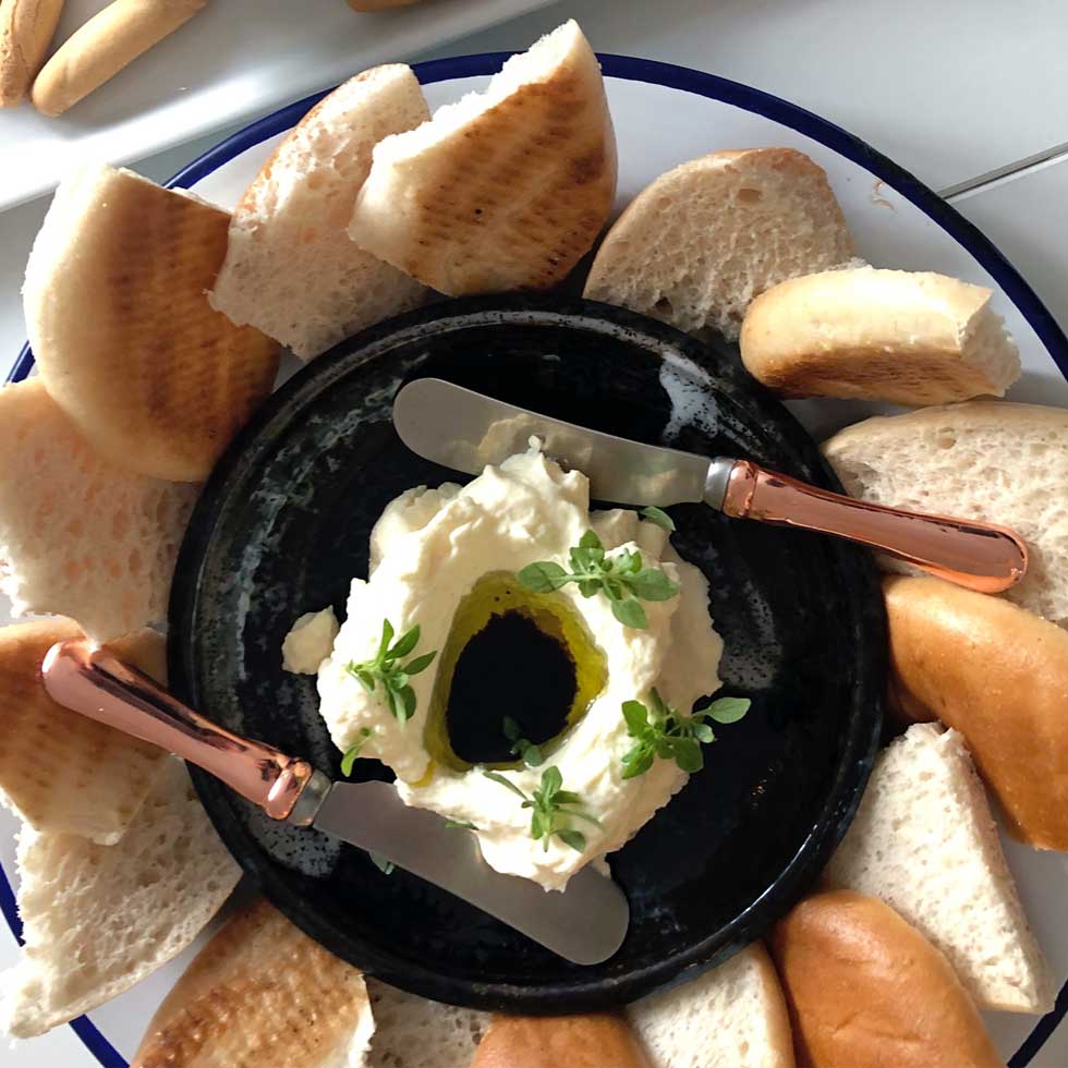 A black plate with a creamy white dip topped with olive oil and herbs, surrounded by slices of pitta bread, and two rose-gold handled spreading knives resting on the plate.