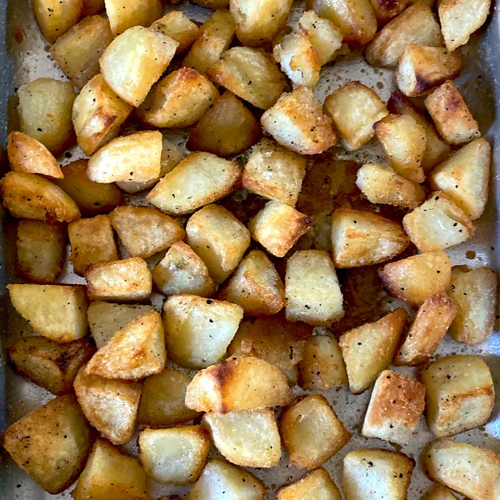 Golden brown roasted potato cubes spread out on a baking tray, seasoned with salt and pepper, showing crispy edges and a slightly oily surface.
