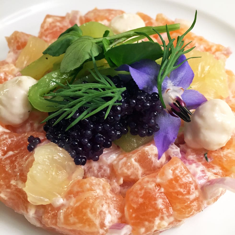 A colourful gourmet salad with orange segments, black caviar, citrus pieces, green herbs, edible flowers, and dollops of creamy sauce, artfully arranged on a white plate.