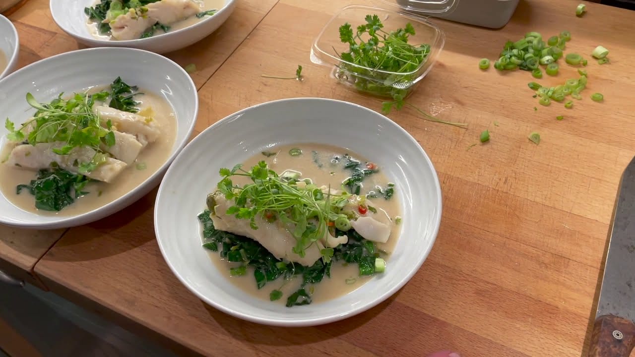 A bowl with fish fillet on a bed of greens, topped with fresh herbs and sauce. Two more similar bowls and a container of herbs are on a wooden worktop with chopped spring onions.
