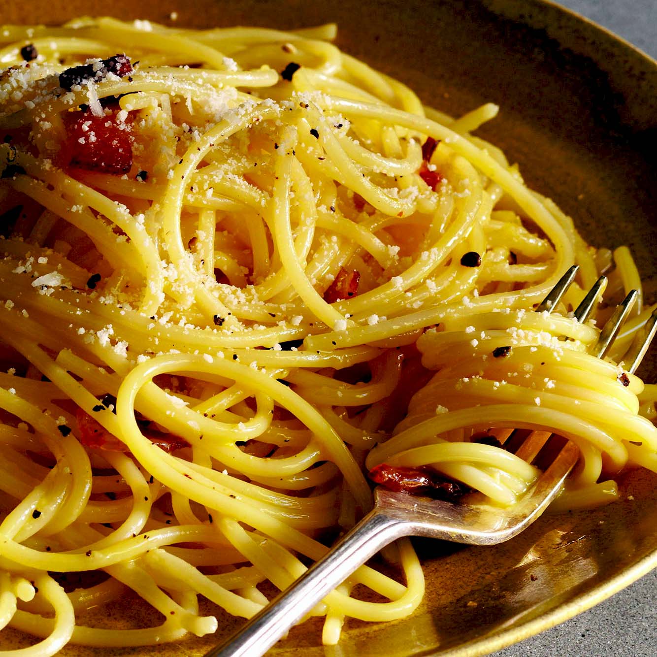 A close-up of cooked spaghetti on a plate, sprinkled with grated cheese and cracked black pepper, with a fork twirling some noodles.