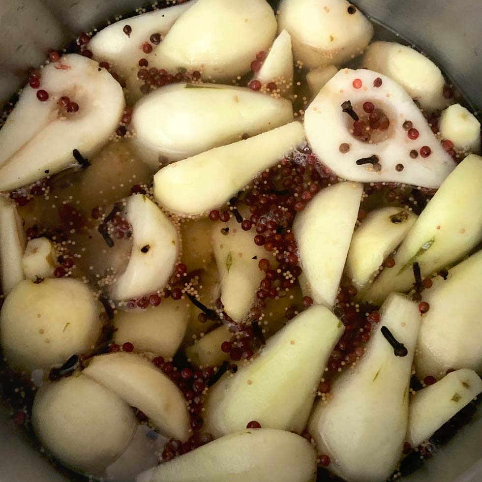 Sliced pears soaking in a saucepan with whole cloves, mustard seeds, and pink peppercorns, possibly being prepared for pickling or poaching.
