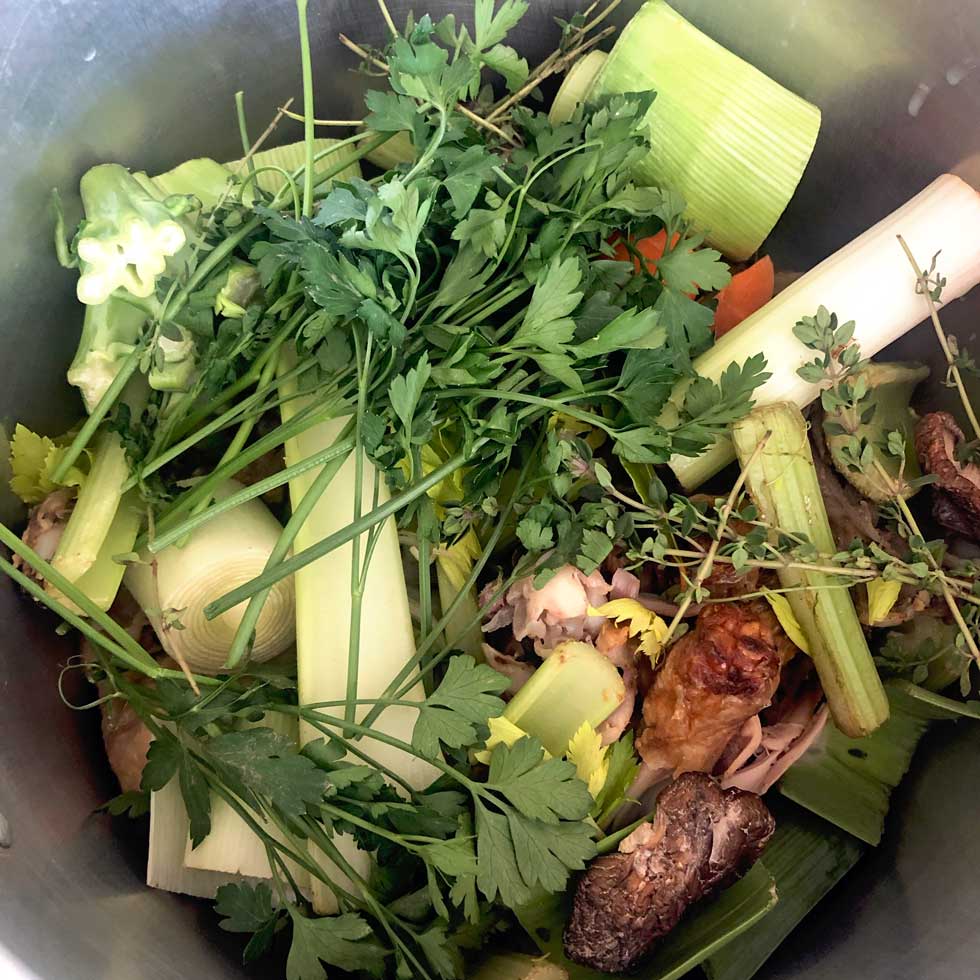 A pot filled with various fresh vegetables and herbs, including leeks, celery, carrots, parsley, thyme, and chives, arranged together and ready for cooking, possibly to make vegetable stock or broth.